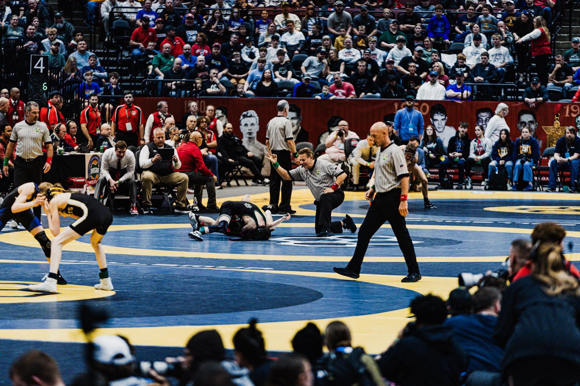 Several wrestling matches under way at once on a multi-mat arena floor, with a packed crowd in the stands